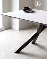 Modern dining table with a white marble top and an asymmetrical matte black metal base, styled with black dishes and clear wine glasses on a neutral-toned room with light wooden flooring.