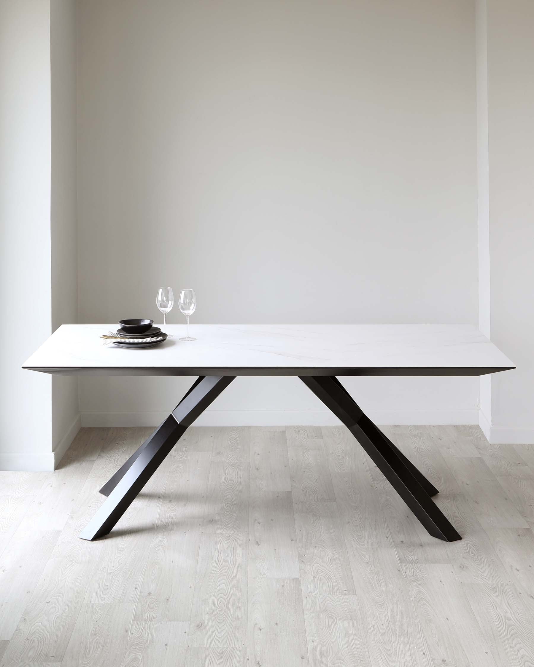 A modern minimalist dining table with a sleek white tabletop and angular black metal legs in an X formation, set against a neutral backdrop with a subtle place setting for two, including black plates and clear wine glasses.