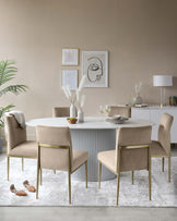 Modern dining room featuring a round white table with a fluted texture design on the base and four contemporary beige upholstered chairs with sleek gold metal legs. In the background, a matching white sideboard, a soft beige wall, and elegant home decor enhance the sophisticated ambiance. A white area rug underlays the dining set.