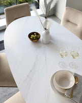 Elegant contemporary dining area featuring an oval white marble table with organic grey veining, complemented by a set of modern minimalist beige upholstered dining chairs with clean lines and subtle curves.