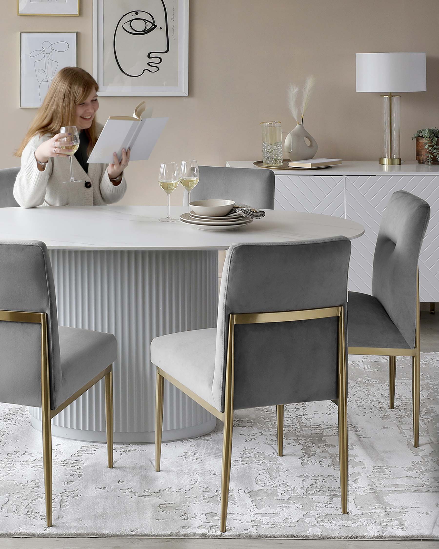 Elegant modern dining room furniture featuring a round white table with a fluted base, paired with luxurious grey upholstered chairs accented by slender gold metal legs. A cream textured area rug lies underneath, complementing the sophisticated neutral palette.