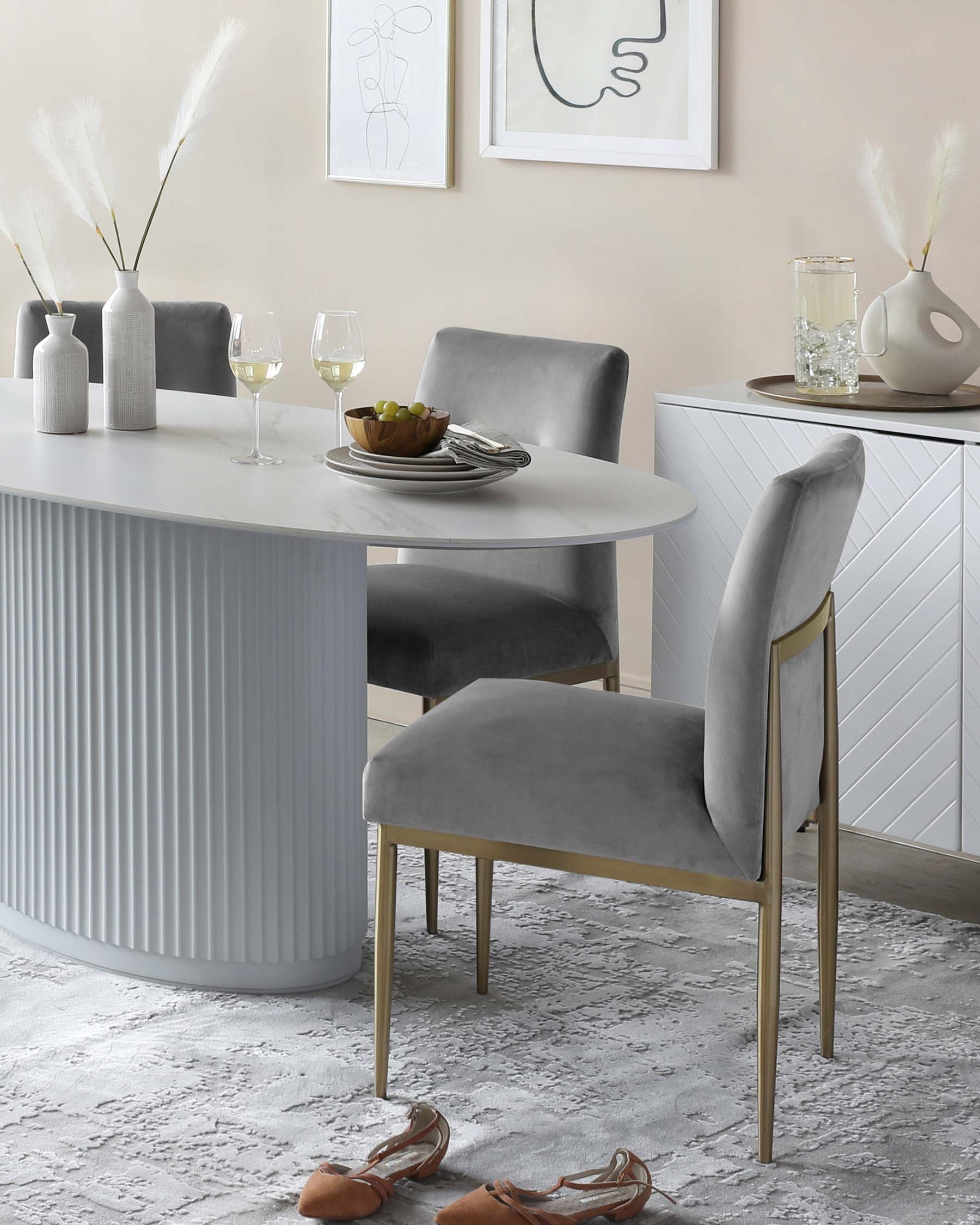 Modern dining room furniture set including a round white table with a fluted vertical panel base and a pair of elegant grey upholstered chairs with slender brass legs, arranged on a textured grey area rug. A matching white and brass sideboard with geometric facades is visible in the background. The scene is accessorized with decorative vases, framed abstract wall art, and a bottle of wine with glasses, imparting a sophisticated ambiance.