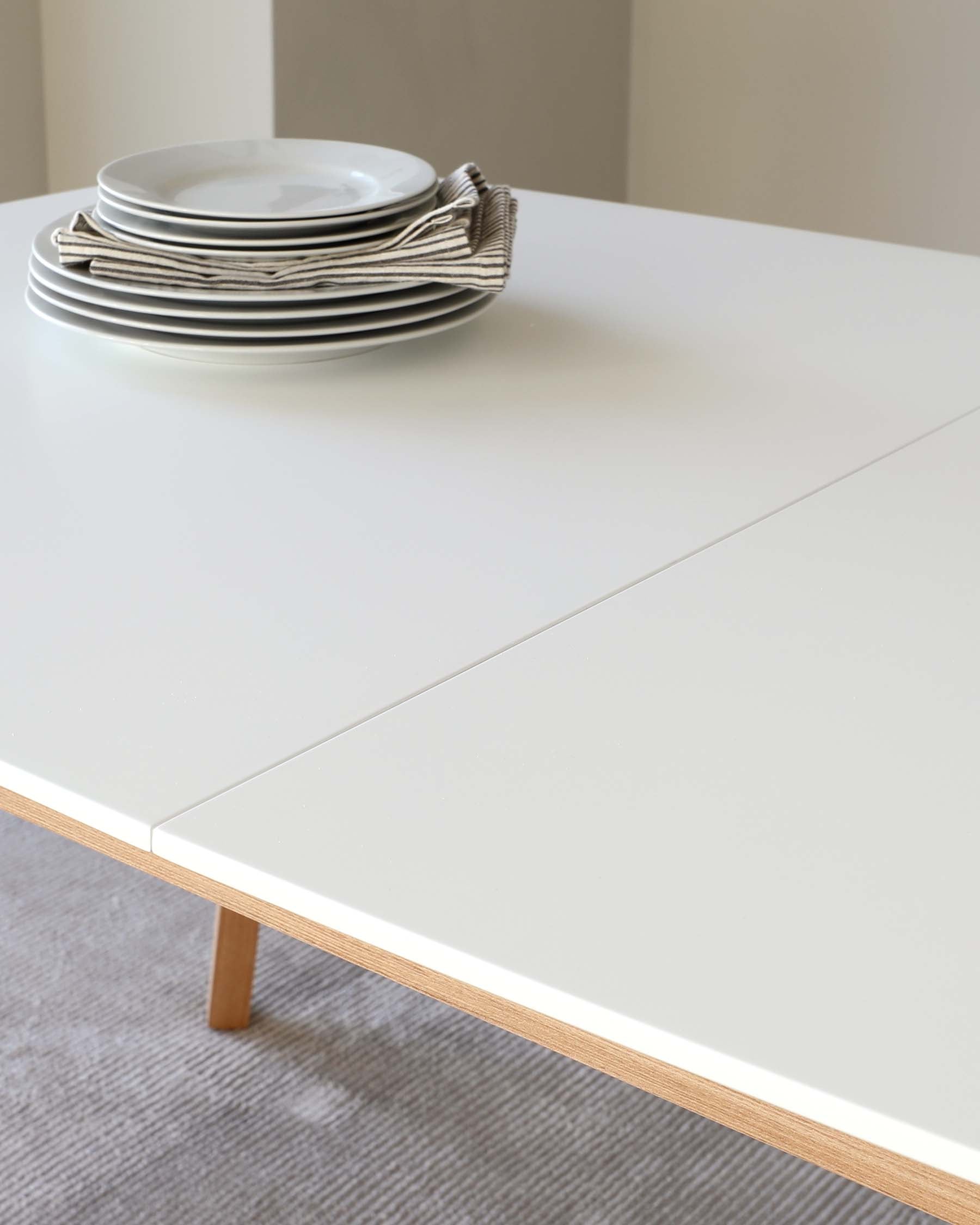 A modern minimalist white dining table with clean lines and natural wood accents on the legs, accompanied by a stack of simple white plates and striped napkins on the surface.