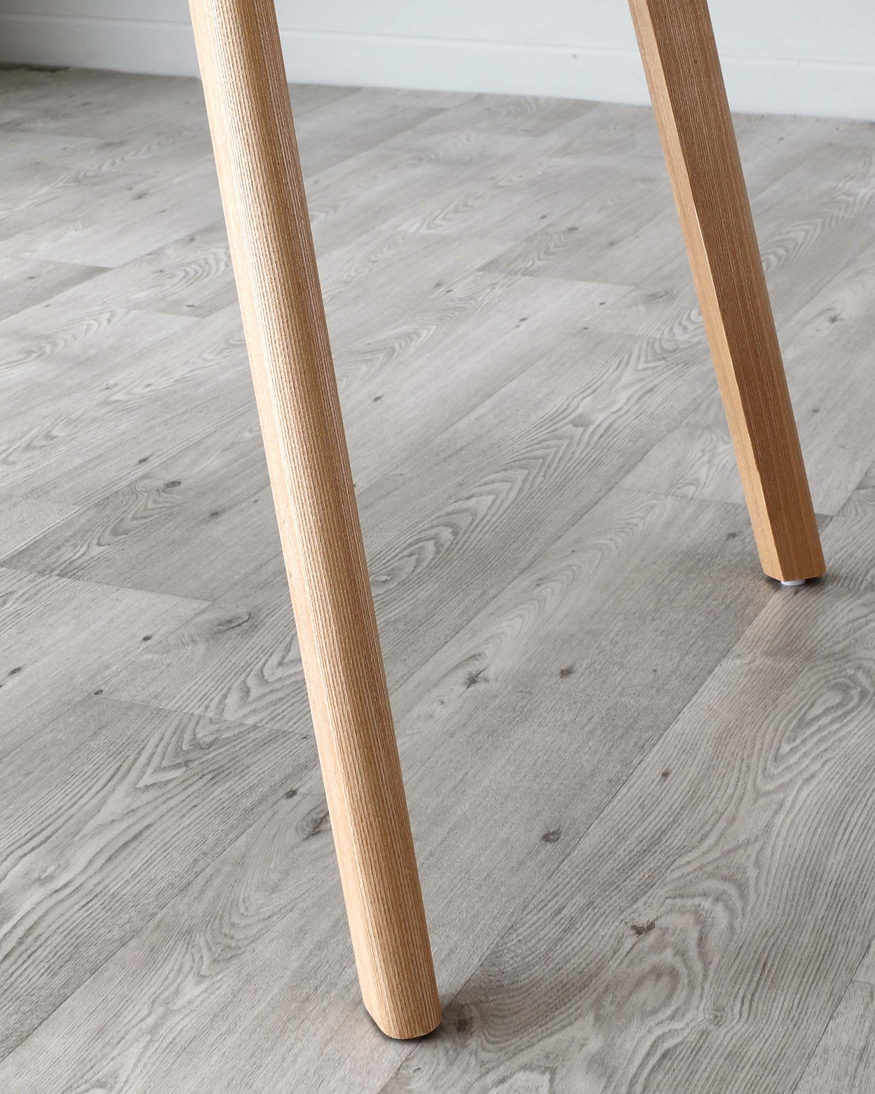 Close-up view of the legs of a wooden table, showing natural wood grain texture, positioned on a grey wood-effect laminate flooring.