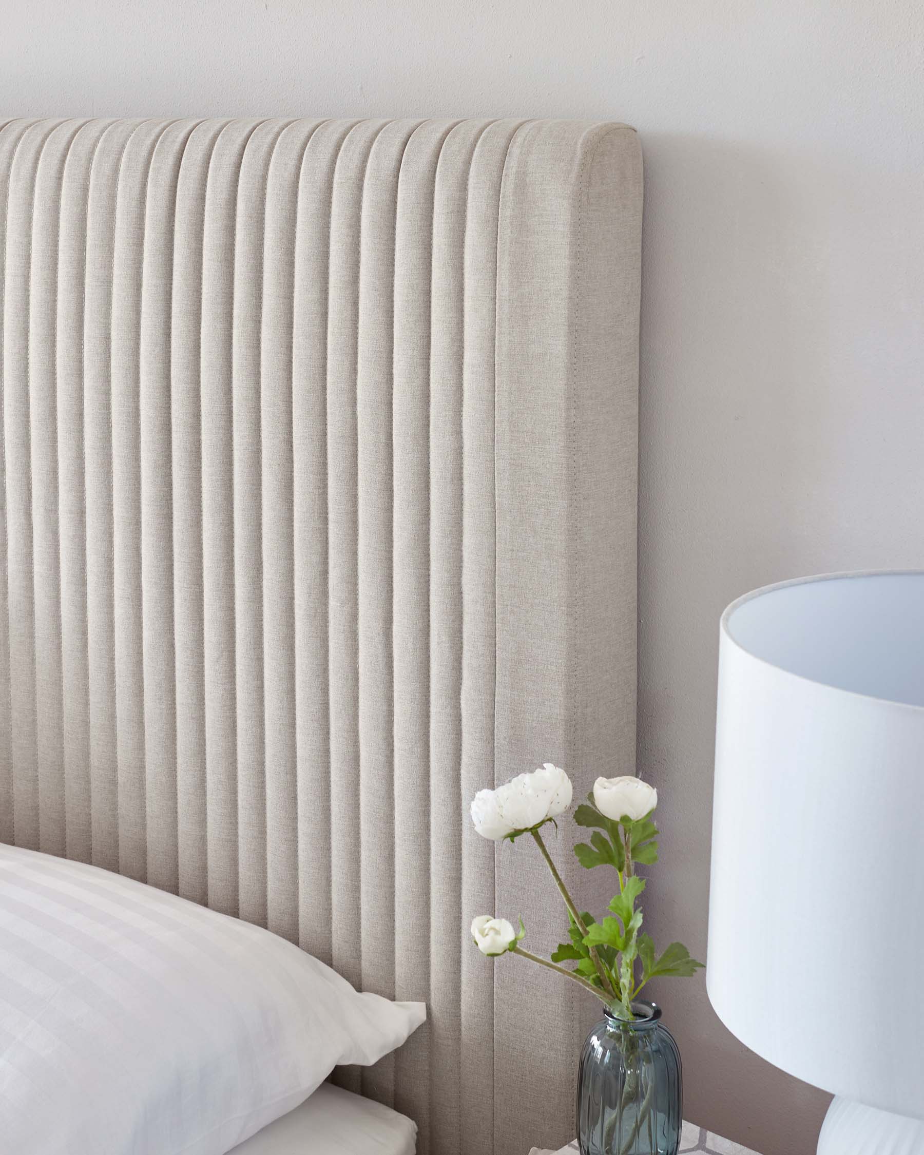 Elegant neutral-toned upholstered headboard with vertical channel tufting, complemented by crisp white bedding and a white lampshade nearby, and accented with a clear vase holding fresh white flowers against a light wall.