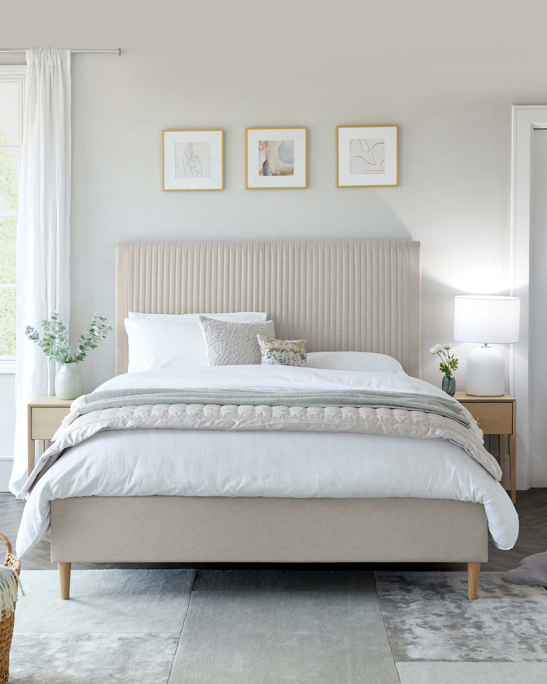 Elegant contemporary bedroom furniture featuring a king-size bed with an upholstered beige headboard displaying vertical channel tufting. The bed is dressed in white linens with a textured grey and white throw and assorted decorative pillows. Flanking the bed are two natural wood nightstands, each with a single white lamp with a classic shade. A plush grey area rug is partially visible beneath the bed.