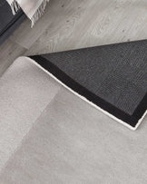 Contemporary two-tone area rug with a light grey section featuring a fringe edge on top of a darker grey bordered section, laid out on a light wooden floor.