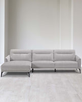 Elegant modern light grey sectional sofa with chaise lounge on the left side, featuring clean lines, button-tufted back cushions, and dark slender legs. The sofa is placed in a minimalist setting with light wooden flooring and a plain white wall background, emphasizing the furniture's contemporary design.