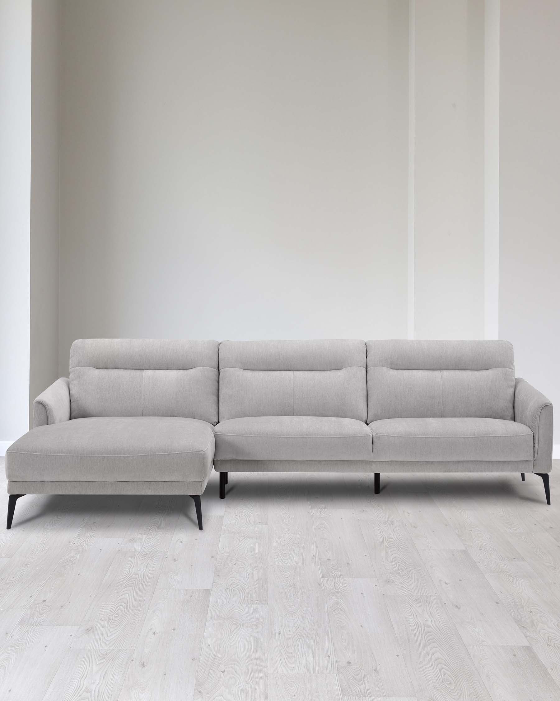 Elegant modern light grey sectional sofa with chaise lounge on the left side, featuring clean lines, button-tufted back cushions, and dark slender legs. The sofa is placed in a minimalist setting with light wooden flooring and a plain white wall background, emphasizing the furniture's contemporary design.