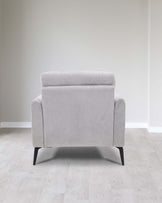 Modern light grey fabric armchair with sleek black wooden legs, positioned on a pale wood floor against a neutral wall.