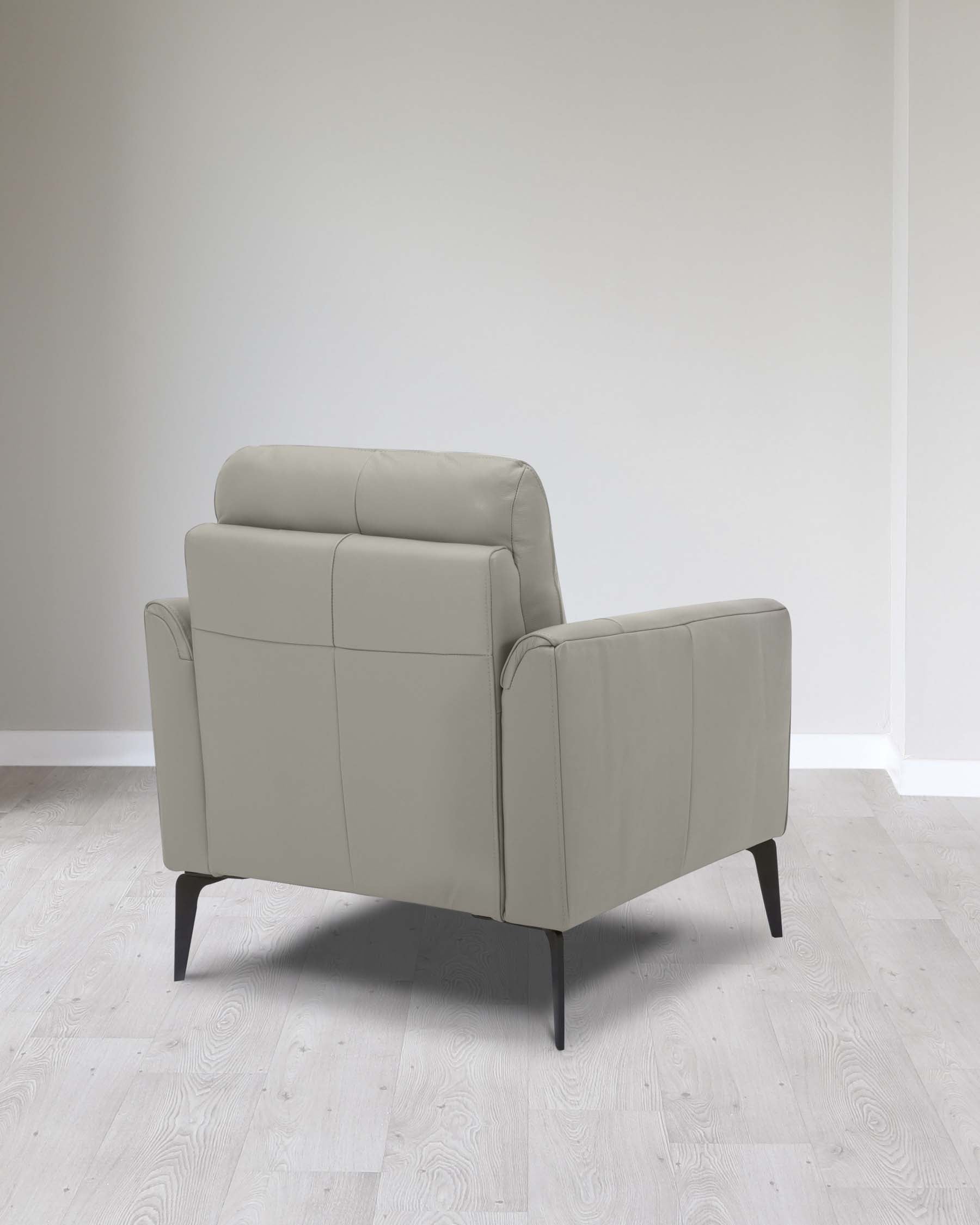 Modern light grey leather armchair with a sleek silhouette, featuring a padded backrest, cushioned seat, and armrests, supported by four dark tapered wooden legs, set against a neutral background with light herringbone-patterned flooring.