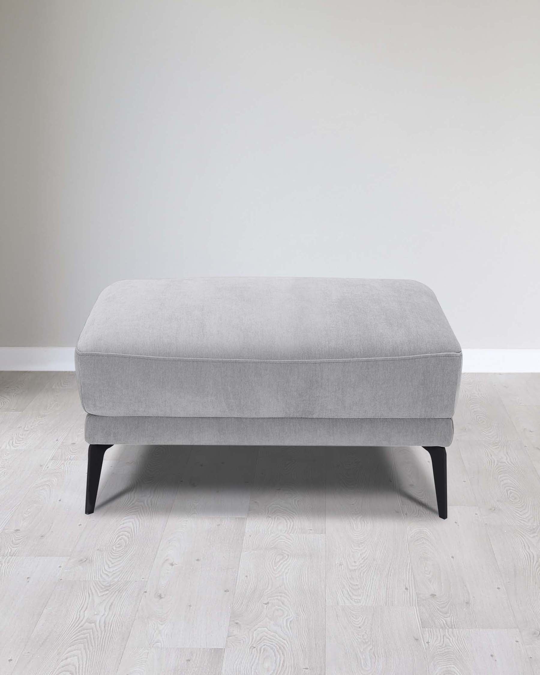 Modern light grey fabric upholstered ottoman with dark tapered wooden legs, showcased against a neutral wall and light wooden floor.