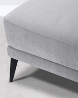 A modern light grey fabric upholstered ottoman with a plush, seamless top cushion and a set of sturdy, angled black wooden legs. The piece is photographed on a white wood floor, highlighting its clean and elegant design.