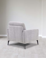 Modern light grey fabric armchair with a cushioned back, boxy structure, and angled black wooden legs set on a light hardwood floor against a neutral wall.