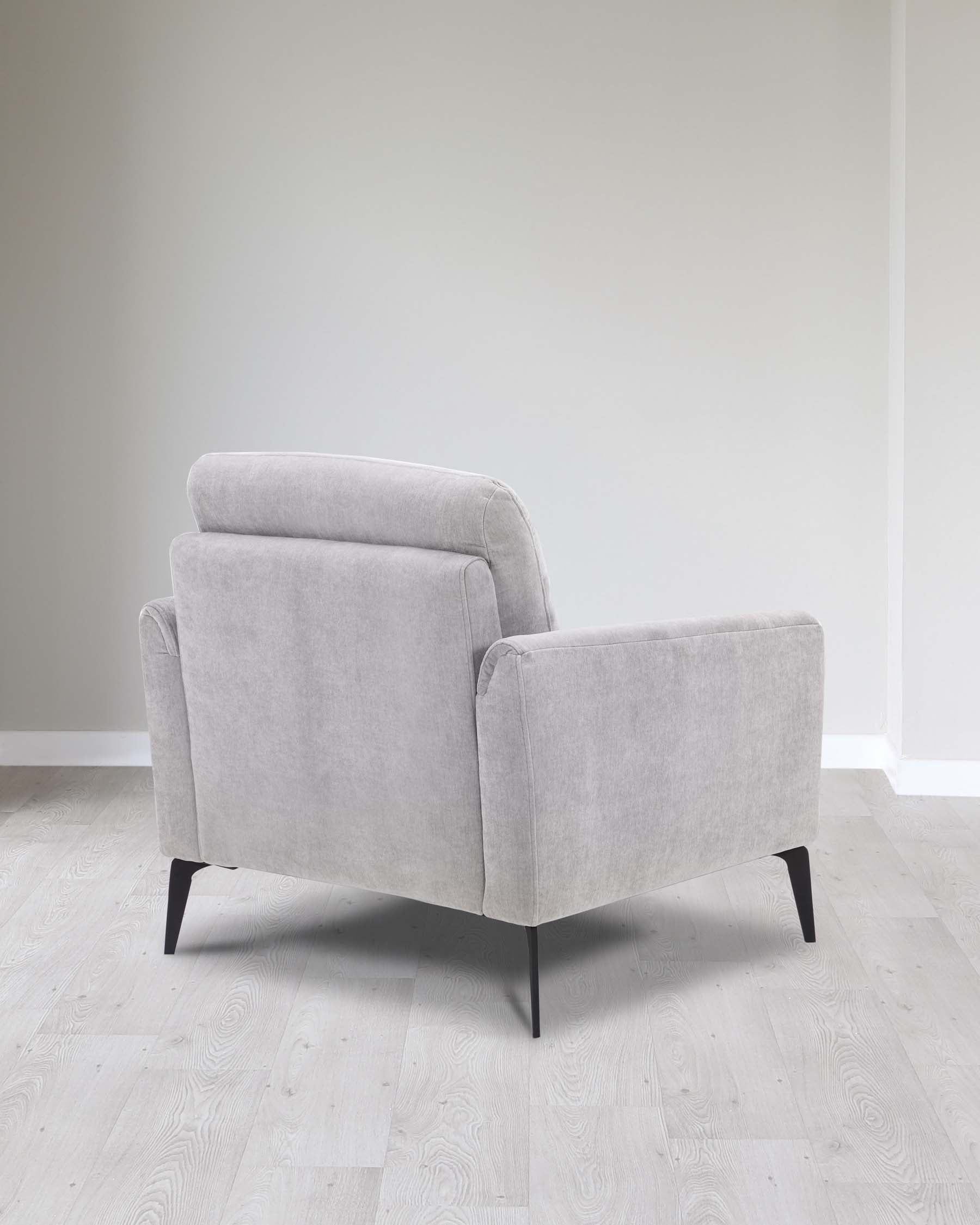 Modern light grey fabric armchair with a cushioned back, boxy structure, and angled black wooden legs set on a light hardwood floor against a neutral wall.