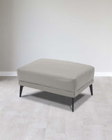 Modern light grey upholstered ottoman with a rectangular cushion top and four black tapered wooden legs, set on a light hardwood floor against a plain white background.