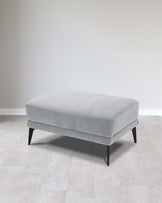 A modern minimalist light grey fabric ottoman with angled black wooden legs, positioned on a light wooden floor against a neutral background.