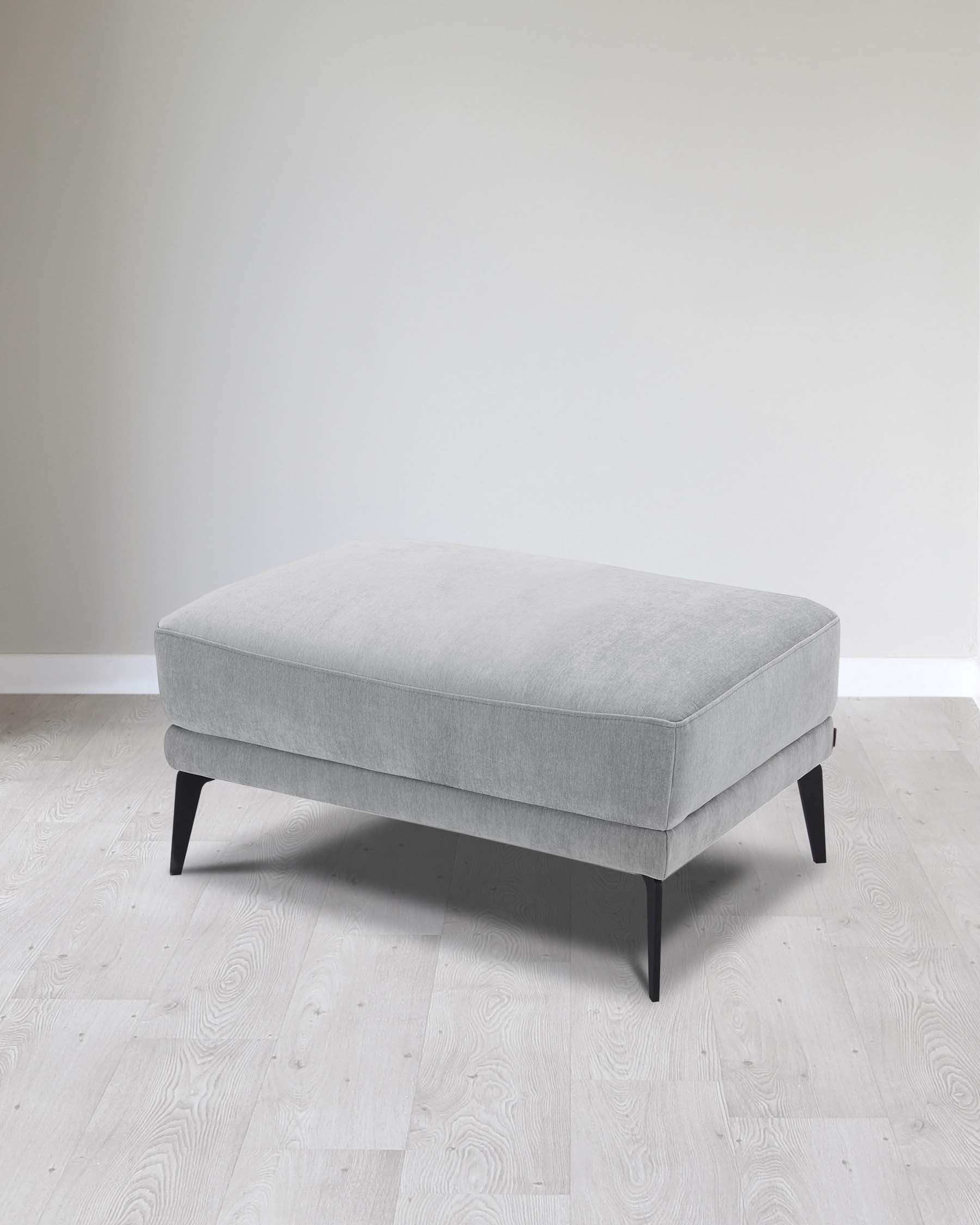 A modern minimalist light grey fabric ottoman with angled black wooden legs, positioned on a light wooden floor against a neutral background.