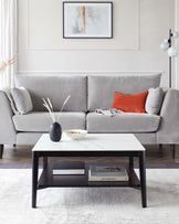 A modern living room with a sleek, minimalist design featuring a light grey upholstered three-seater sofa with plush cushions, accompanied by a rectangular white marble-top coffee table with a black frame and a lower shelf displaying books. The room is accented with a vibrant orange throw pillow, a pair of glasses, and a decorative vase with dried pampas grass on the coffee table.