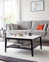 A modern living room featuring a light grey upholstered three-seater sofa with plush cushions and a flannel throw draped over its arm, complemented by a stylish rectangular coffee table with a black frame and white marble top, set upon a grey and white area rug.