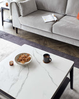 Contemporary living room with a light grey upholstered sofa and a white marble-top coffee table with black legs. The table is accessorized with a decorative bowl, candles, and a cup.