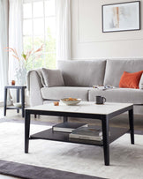 A contemporary living room setting featuring a light grey upholstered three-seater sofa with a textured throw blanket and an orange decorative pillow. In front of the sofa is a rectangular black coffee table with a white top, holding books, a mug, and a small bowl. To the left, there is a small round side table in a dark colour with a vase and coaster on top. The room is completed by a soft grey area rug, sheer white curtains, and a framed abstract art piece on the wall.