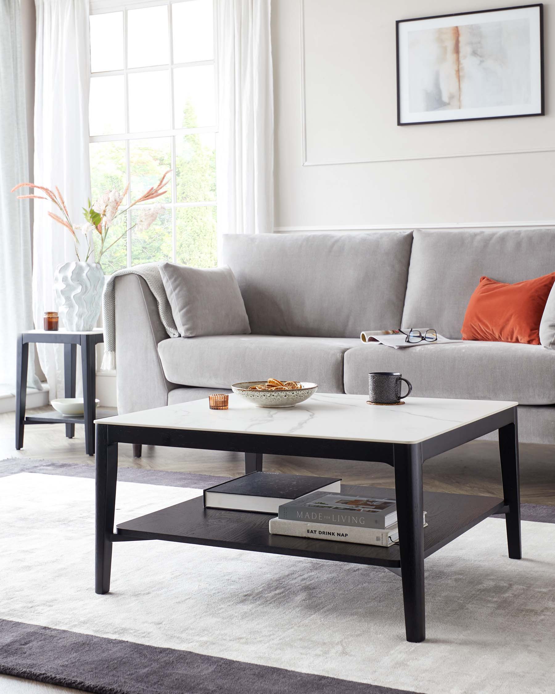 A contemporary living room setting featuring a light grey upholstered three-seater sofa with a textured throw blanket and an orange decorative pillow. In front of the sofa is a rectangular black coffee table with a white top, holding books, a mug, and a small bowl. To the left, there is a small round side table in a dark colour with a vase and coaster on top. The room is completed by a soft grey area rug, sheer white curtains, and a framed abstract art piece on the wall.