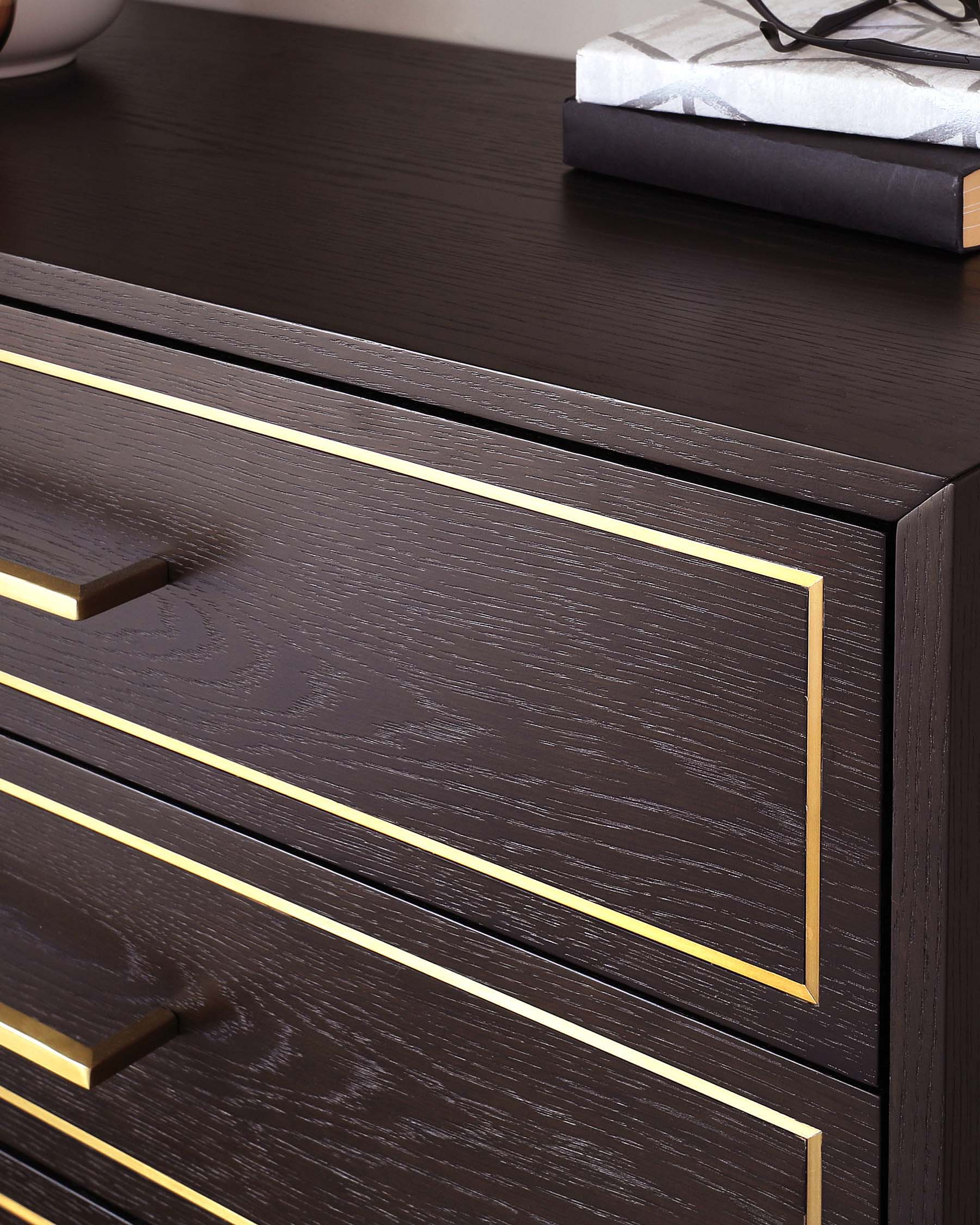 Elegant contemporary dark wood dresser featuring clean lines, with brass-coloured metal trim and handles accentuating the front of its drawers. A book with a black spine and grey cover is placed on its smooth, polished surface.