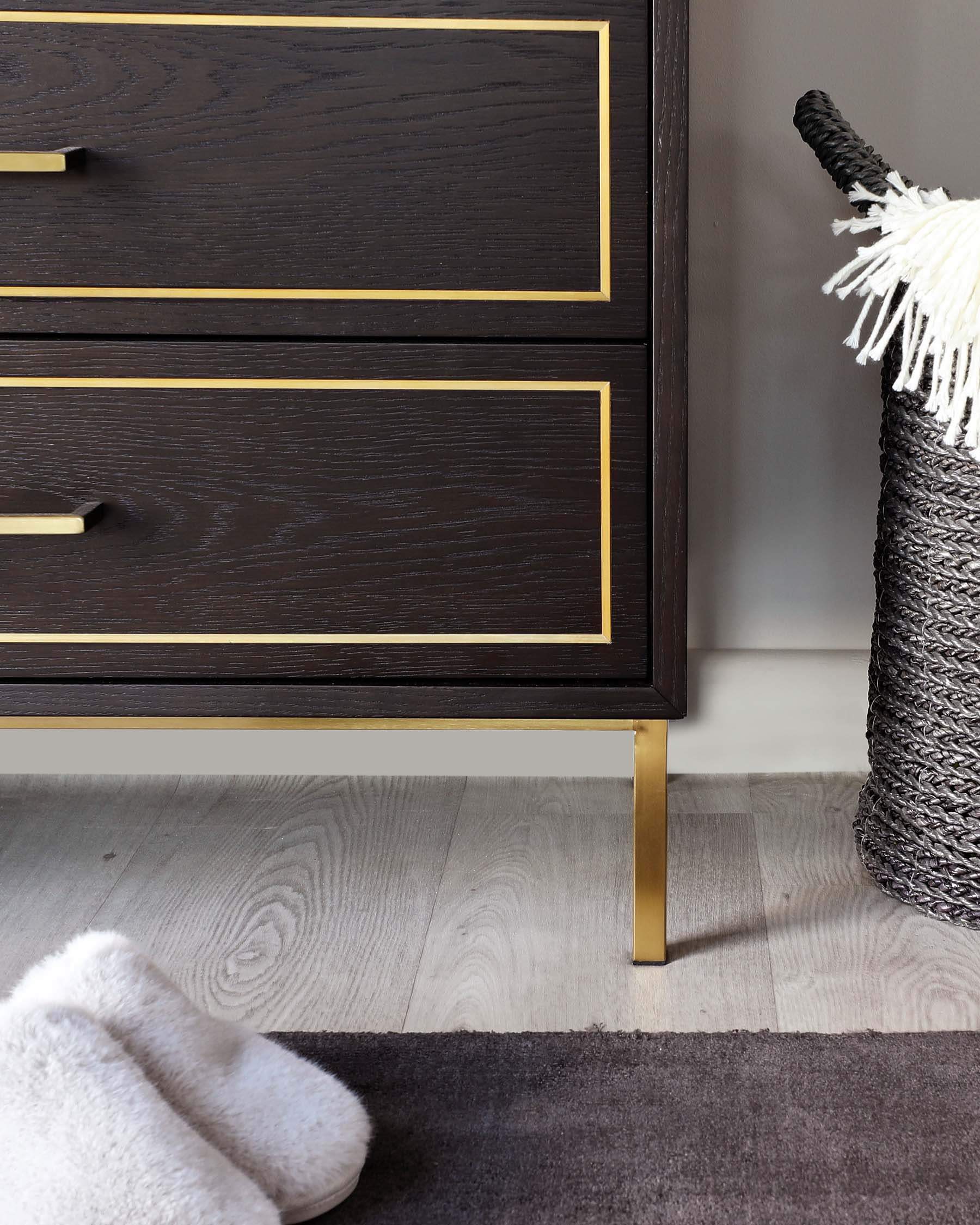 Elegant dark wood chest of drawers with brass-coloured metal accents and handles, displayed in a modern interior setting next to a woven basket and fluffy white slippers on a dual-textured rug.