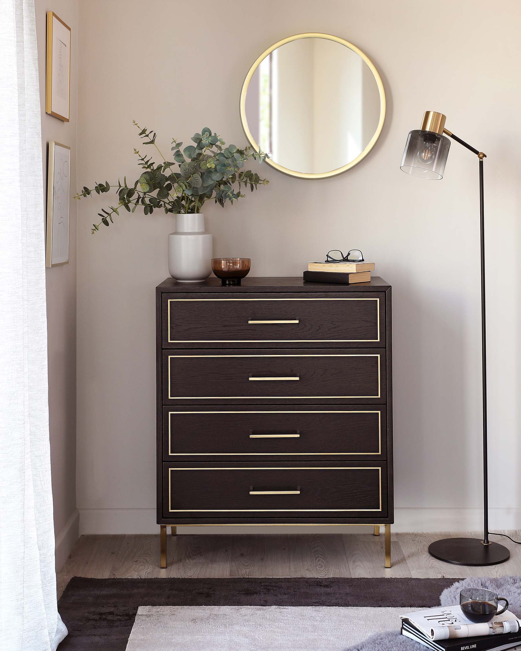 A modern dark-toned wooden dresser with four drawers, featuring brass-finished handles and legs. A decorative vase with green foliage, a small brown bowl, and a pair of eyeglasses resting on a book are placed on top, complementing the aesthetics. In close proximity, a sleek floor lamp with a brass accent and a clear glass shade provides ambient lighting. The setting is further enhanced by a circular mirror with a thin brass frame on the wall above the dresser.