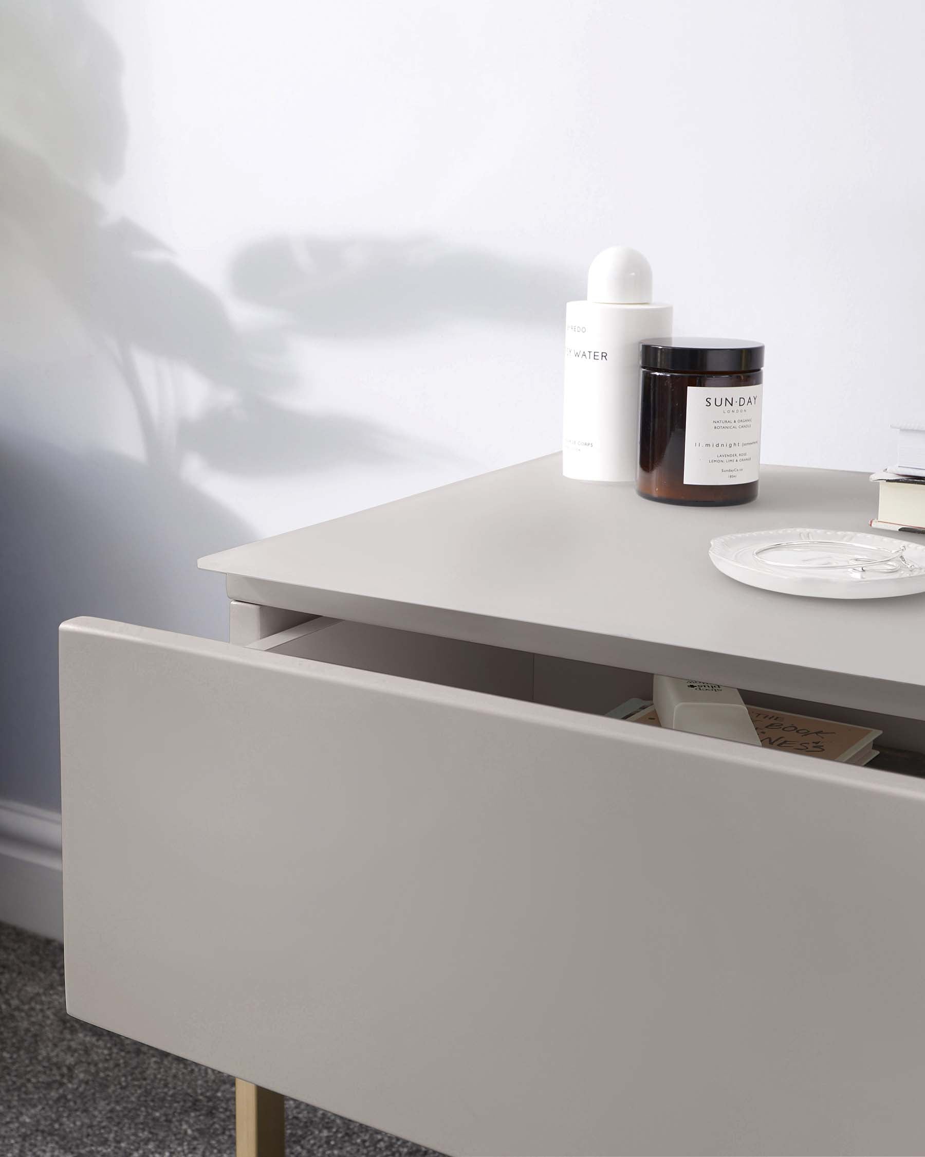 Modern minimalist light beige side table with a sleek surface, featuring an open drawer revealing books and other items. The table is supported by four slender, angled wooden legs. On top of the table, there is a curated arrangement of two cylindrical containers labelled 'WATER' and 'SUNDAY' and a small ornate plate. The background suggests a bright, softly lit room.