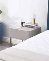 Modern minimalist-style bedside table in a muted grey finish with a single drawer and slender gold legs.