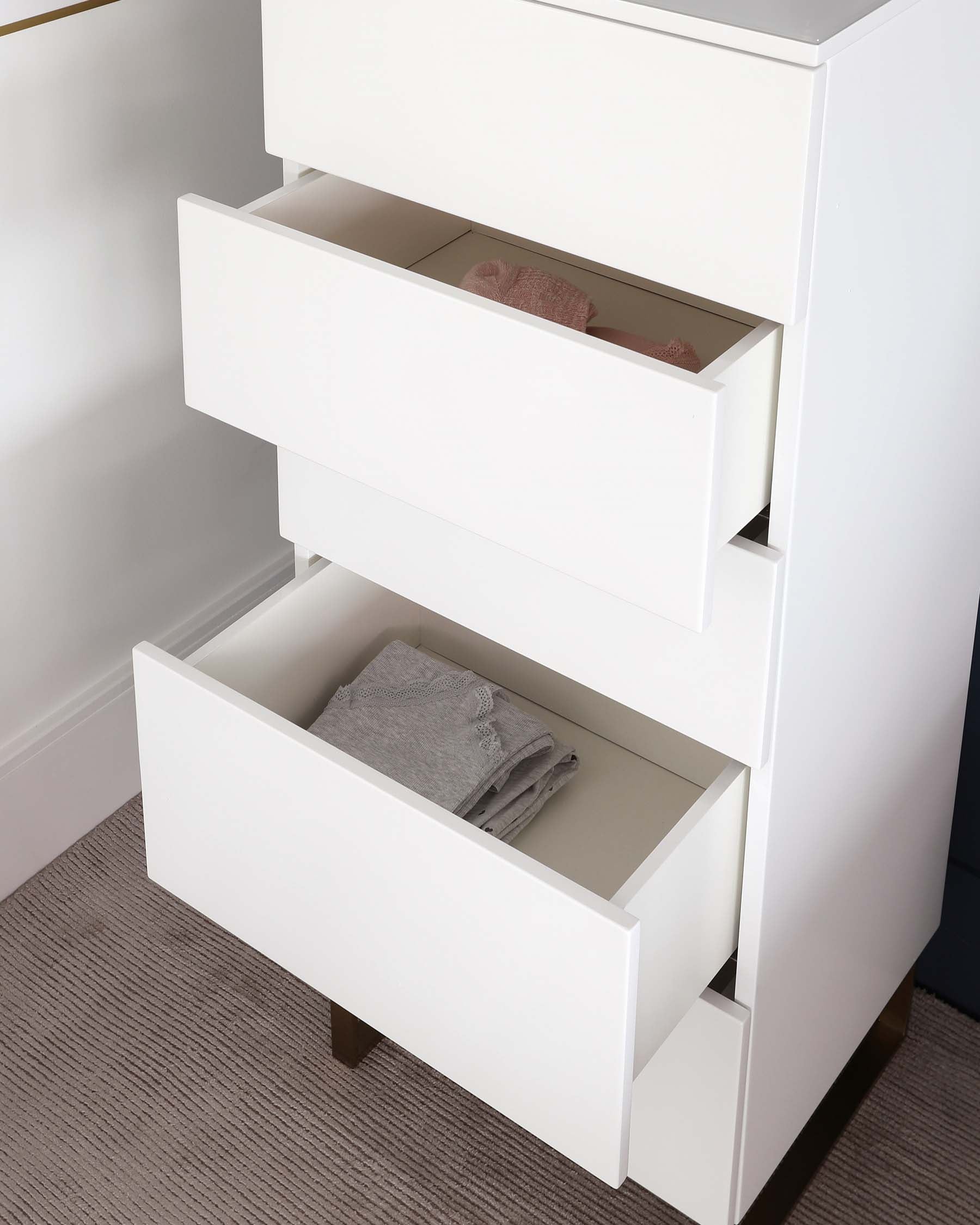 A modern white chest of drawers with three tiers. The top two drawers are partially open, displaying neatly folded towels within. The unit has a sleek, minimalist design with no visible handles, standing on a low-profile base.