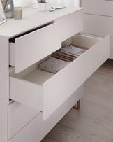 A modern white dresser with an open drawer revealing neatly folded blankets, featuring sleek handles and minimalist wooden legs. The dresser is accessorized with a few decorative items, including a candle, succulent plant, and picture frame.