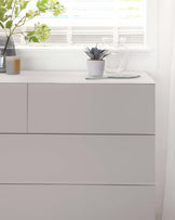 Modern minimalist light grey three-drawer dresser with a smooth finish and no visible hardware, placed against a white wall near a window with white venetian blinds. Decorated with a vase of greenery, a small potted plant, and a clear glass pitcher on top.