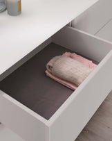 A modern white dresser drawer partially opened, revealing a neatly organized interior with a dark grey lining and a stack of soft-textured pale pink textiles, suggesting a warm, minimalist design suitable for contemporary home decor.