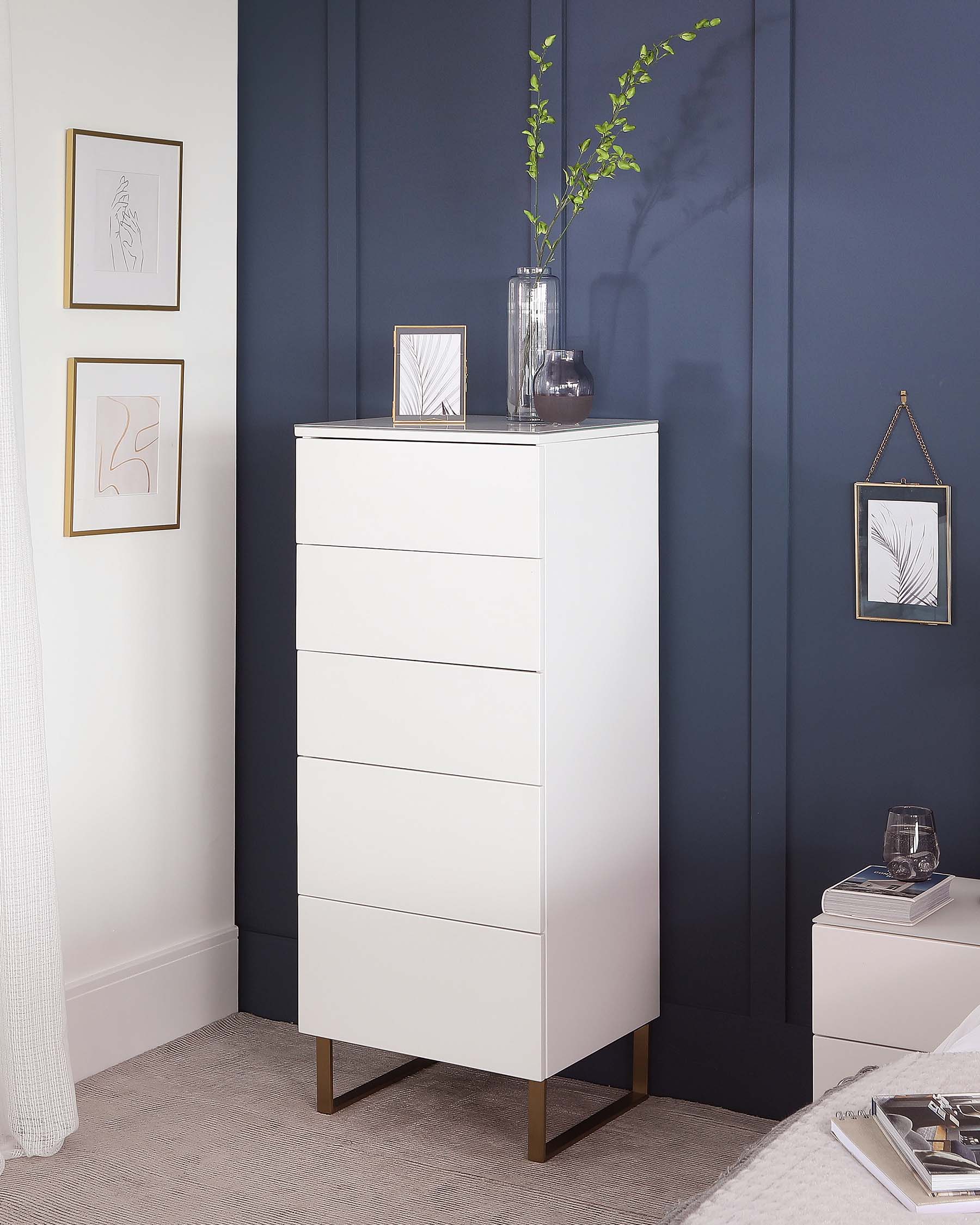 White modern tallboy chest of drawers with brass-finished metal legs, featuring five drawers with flush handles and a sleek, minimalist design. The furniture piece is complemented by a clear glass vase with greenery atop and is set against a dark blue wall adorned with framed artwork, creating an elegant and contemporary interior scene.