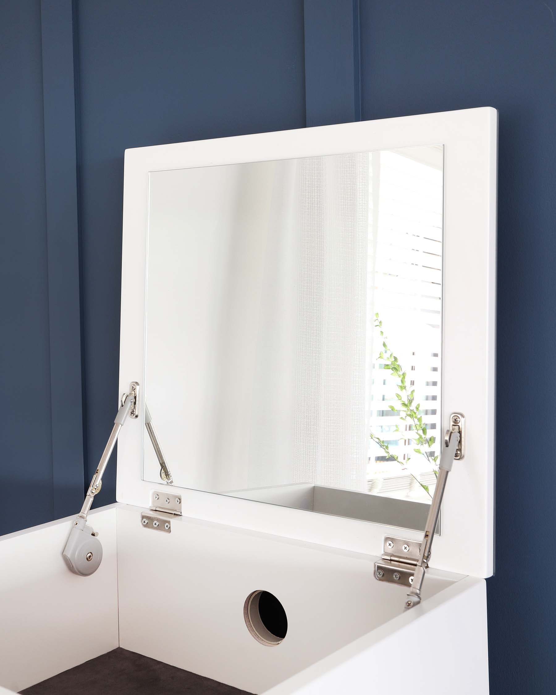 Modern white vanity desk with a large square mirror and metal lifting hinge mechanisms. Visible are two open flaps on the desk revealing interior storage space, and a round cable management hole on the right side of the desk. The furniture is set against a blue wall with a white curtain partially reflected in the mirror.