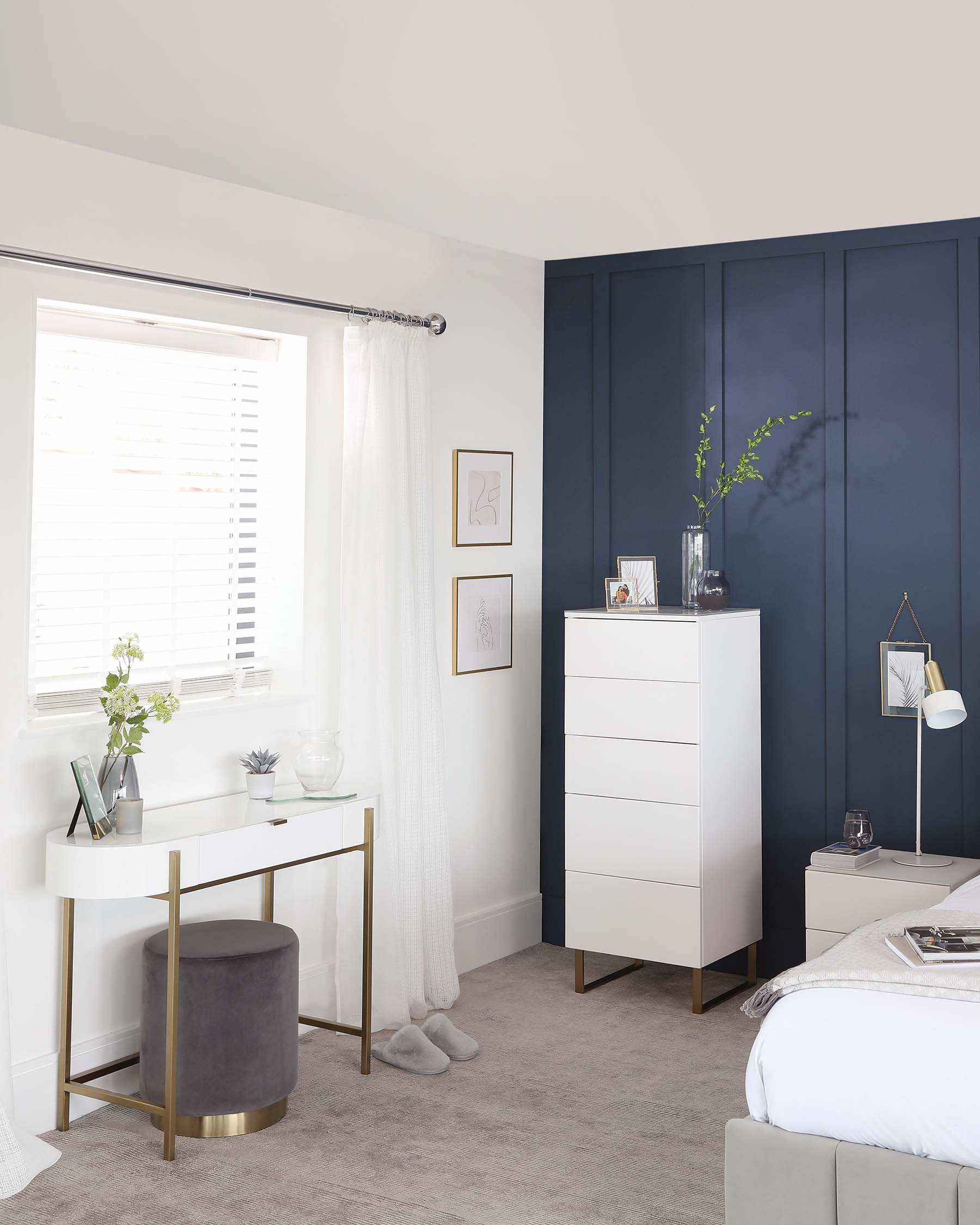 An elegant bedroom scene showcasing modern minimalist furniture including a white five-drawer tall dresser with subtle metal handles and slim wooden legs, a sleek white nightstand with two drawers and slim metal handles, and a white desk with a single drawer accented by a gold frame and handle, paired with a round grey upholstered stool with a matching gold base.