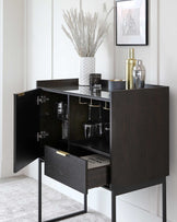 Contemporary black wood bar cabinet, featuring a rectangular silhouette with a dark finish, mounted on a sleek, black metal frame. Two doors, one drawer, and visible shelving are open to reveal interior storage with compartments for glassware and bottles, accented by gold-toned hardware.