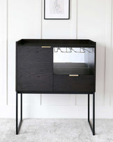 Modern black wooden bar cabinet with glassware shelf, featuring a streamlined design with two drawers and open shelving, elevated on slender metal legs.