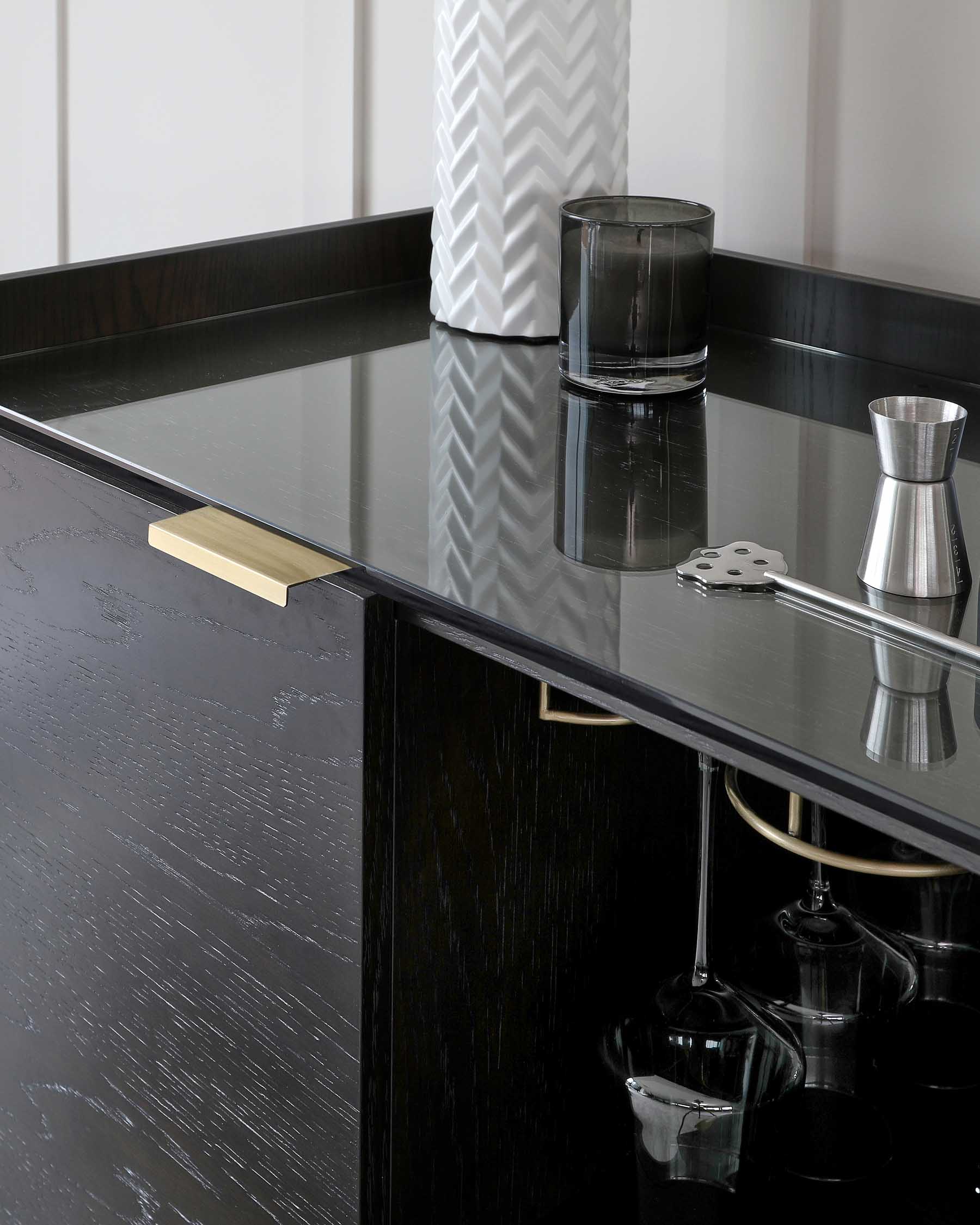 Elegant black bar cabinet with a glossy finish and brushed brass hardware, featuring a glass top surface and integrated wine glass holders beneath the overhanging counter.