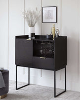 Modern black cabinet with a sleek, minimalist design, featuring a pair of storage compartments, one open shelf with wine glass holders, and a slim drawer with a brass-coloured handle. The cabinet is elevated on slender, straight black metal legs.