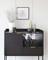 Modern black sideboard with a glossy finish featuring two concealed drawers with a gold handle on one and an open shelf below with glassware on display. The sideboard is set against a white wall with decorative items on top, including a framed line drawing, a vase with white branches, and assorted metallic bar accessories.