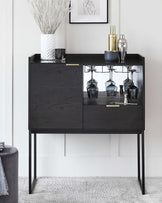 Modern black bar cabinet with elegant brass hardware, featuring a built-in wine glass rack under an open shelf, topped with a solid surface for display, and enclosed storage space below. The unit stands on slender metal legs, offering a sophisticated and contemporary aesthetic.