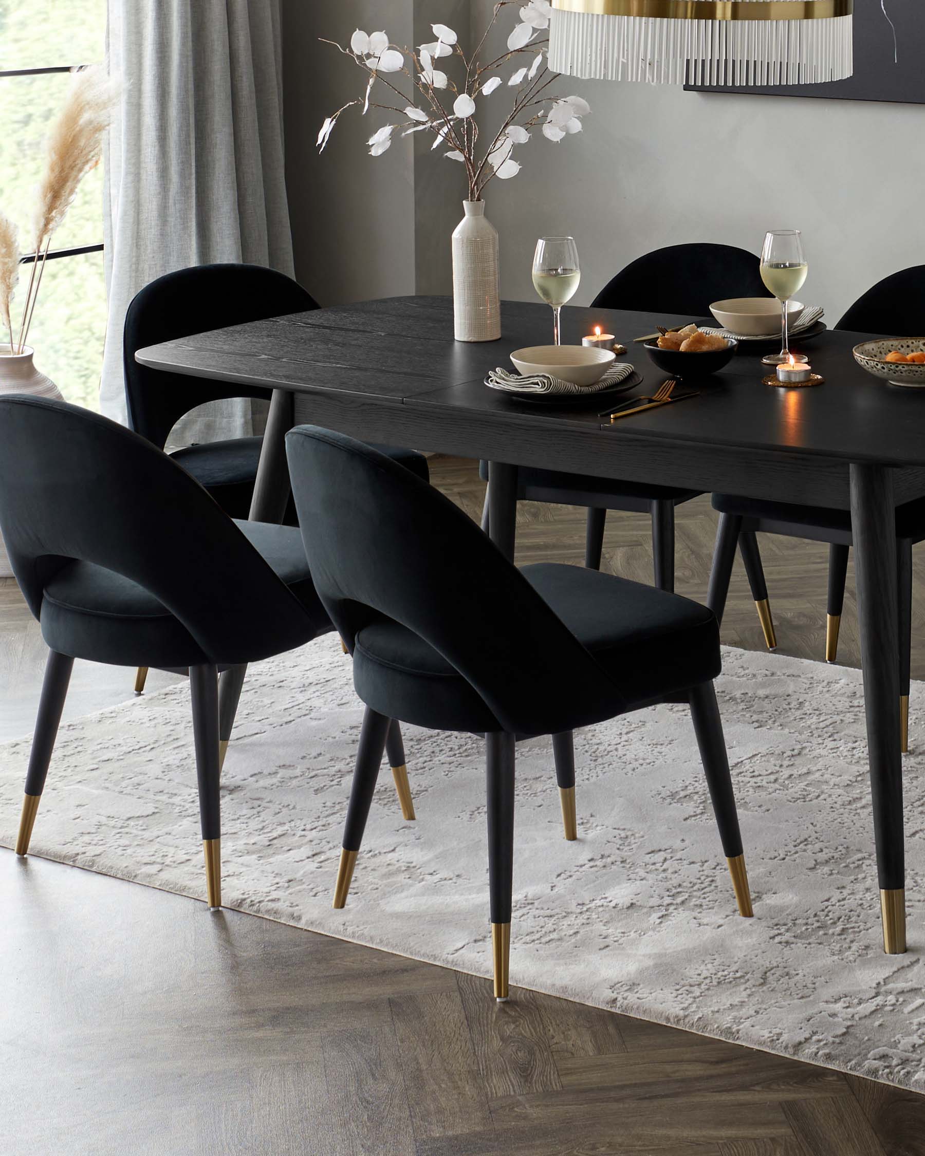 Elegant modern dining furniture set featuring a minimalist matte black rectangular table with distinctive angled legs and a set of four luxurious black upholstered dining chairs with curved backs and slim legs finished in black and accented with golden feet tips, all arranged on a textured light grey area rug.