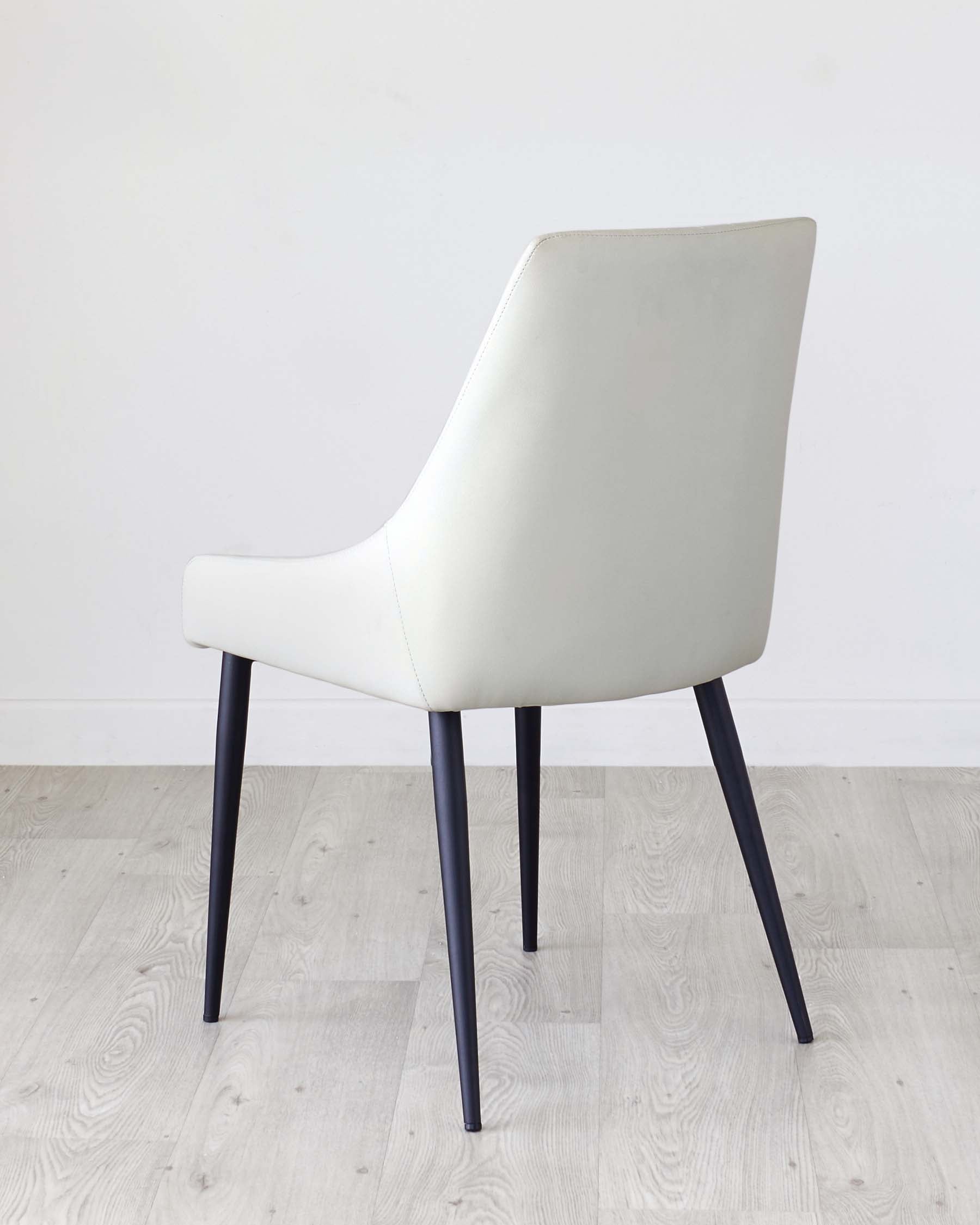 Modern minimalist dining chair with a sleek white leather upholstery and contrasting dark wooden legs set against a light wooden floor and plain white background.