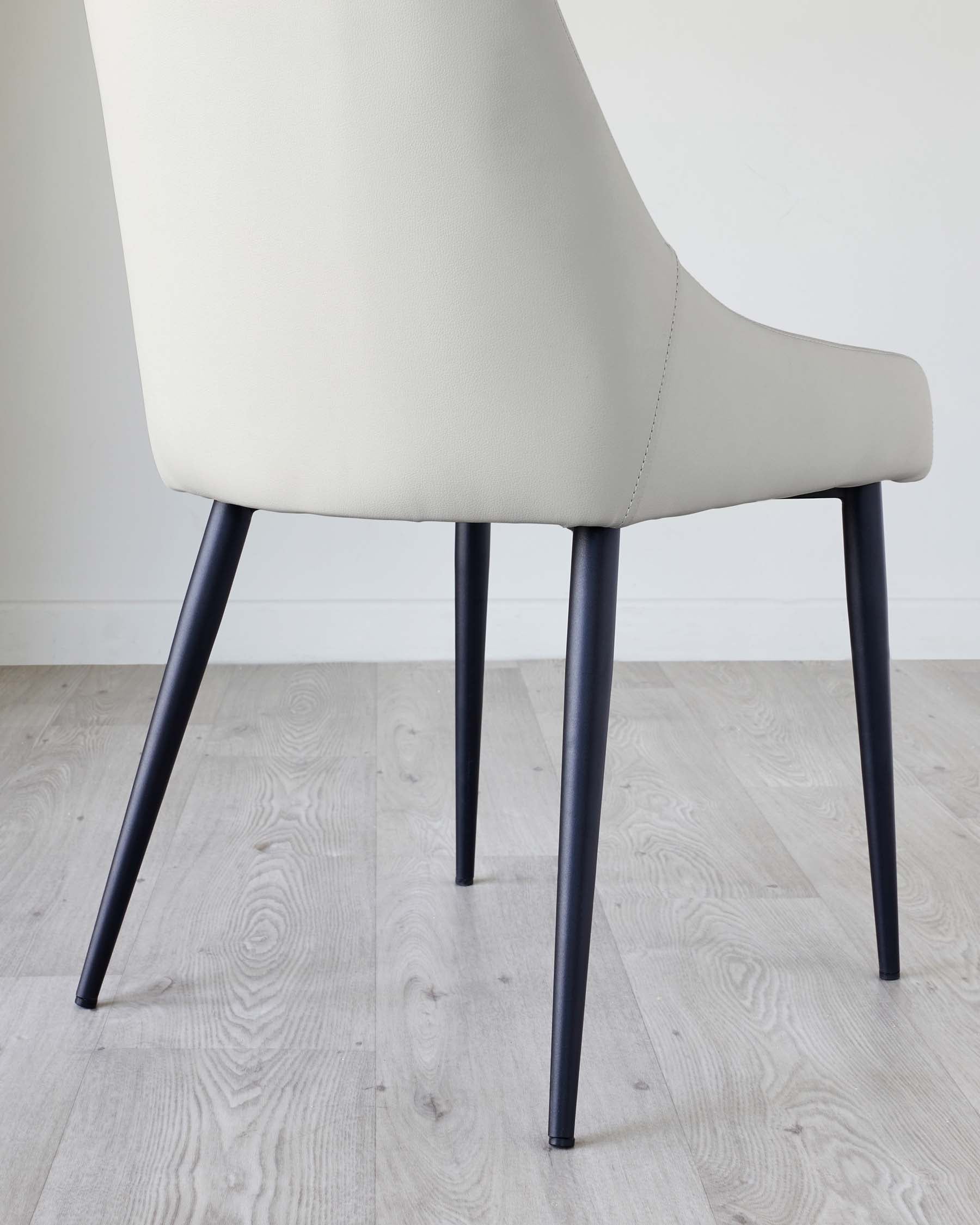 Elegant modern dining chair with a beige upholstered seat and backrest, featuring sleek dark legs that taper slightly towards the floor, set against a light wooden floor with a white wall background.