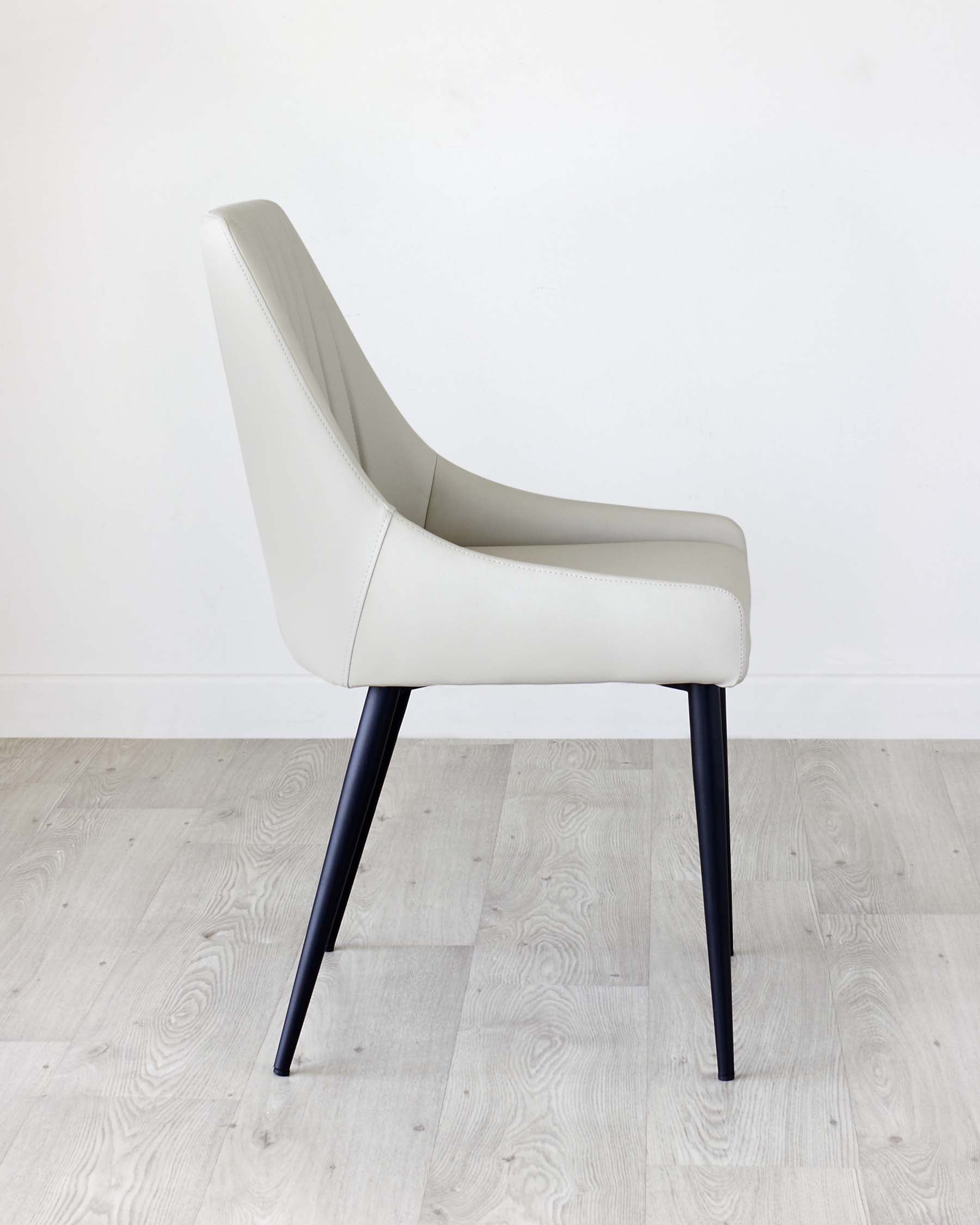 Modern light grey upholstered dining chair with a curved backrest and sleek black metal legs, showcased on a light wood floor against a white backdrop.