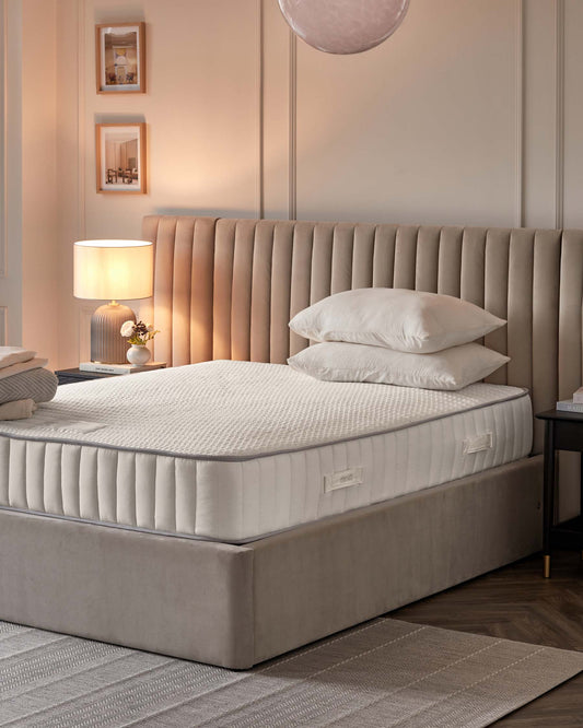 Elegant bedroom featuring a plush king-size bed with an oversized, vertical channel-tufted headboard in a soothing beige fabric. The bed is complemented by a thick, quilted mattress and crisp white bedding. A contemporary nightstand with a dark finish and brass accents is visible at the side, topped with a white lamp boasting a translucent glass base and a warm-toned shade.